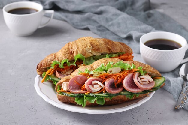 Dos croissants-sándwiches con lechuga, zanahorias, pepinos, salchichas, queso y dos tazas de café sobre una mesa de hormigón gris claro. Concepto de desayuno o almuerzo. Formato horizontal