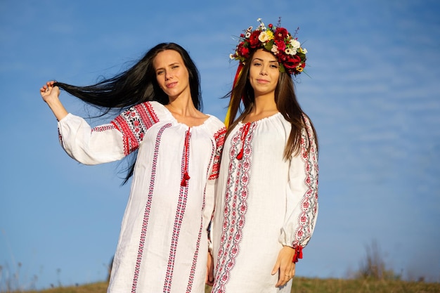 Dos damas bastante jóvenes visten ropa tradicional ucraniana y flores caminan en el campo de trigo hermosa chica étnica en una corona floral decorada a mano admiran la naturaleza Puesta de sol