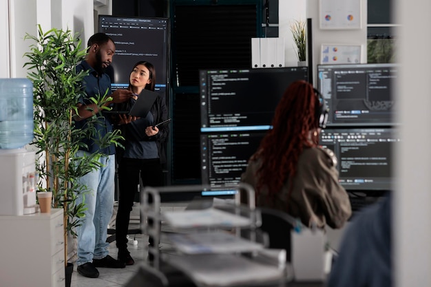 Dos desarrolladores de aplicaciones analizan el código html en busca de errores en la computadora portátil, revisando el gran monitor de televisión en la oficina de la agencia. Codificadores que trabajan en la innovación de seguridad cibernética en una empresa de software ocupada.