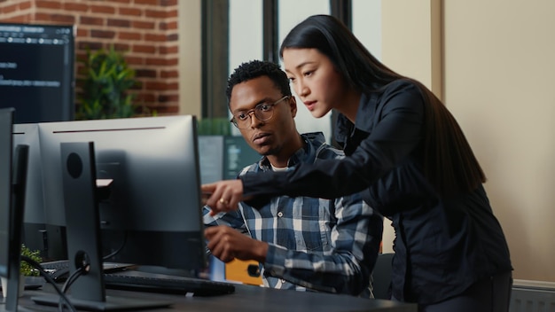 Dos desarrolladores de software que vienen al escritorio y se sientan sosteniendo una computadora portátil con interfaz de codificación y apuntando a la pantalla de la computadora. Equipo de programadores discutiendo algoritmos mirando monitores con código.