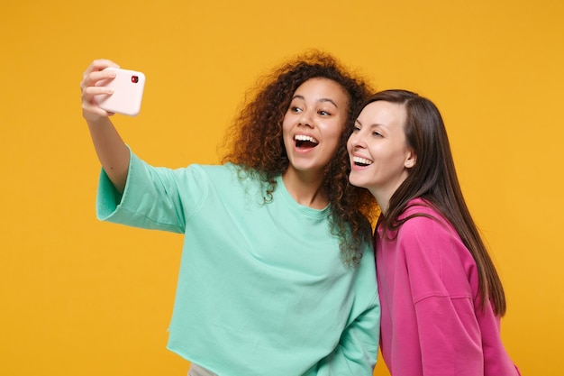 Dos divertidas amigas europeas y afroamericanas vestidas de verde rosa posando aisladas en un fondo amarillo. Concepto de estilo de vida de las personas. Simulacros de espacio de copia. Haciendo autofoto en el teléfono móvil.