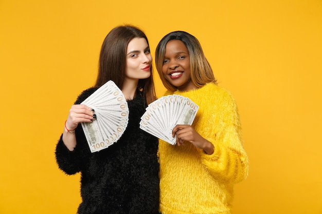 Dos divertidas amigas jóvenes europeas y africanas vestidas de amarillo negro sosteniendo un abanico de dinero en efectivo en billetes de dólar aislados en el fondo de la pared naranja. Concepto de estilo de vida de las personas. Simulacros de espacio de copia.