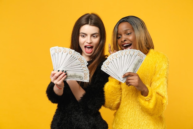 Dos divertidas amigas jóvenes europeas y africanas vestidas de amarillo negro sosteniendo un abanico de dinero en efectivo en billetes de dólar aislados en el fondo de la pared naranja. Concepto de estilo de vida de las personas. Simulacros de espacio de copia.