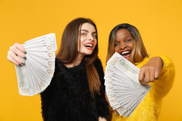 Dos divertidas amigas jóvenes europeas y africanas vestidas de amarillo negro sosteniendo un abanico de dinero en efectivo en billetes de dólar aislados en el fondo de la pared naranja. Concepto de estilo de vida de las personas. Simulacros de espacio de copia.