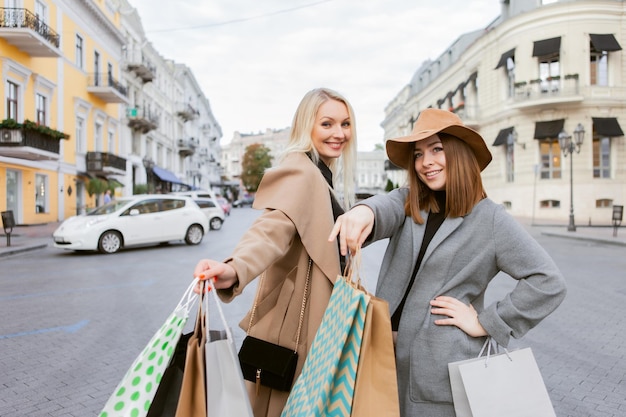 Dos divertidas novias de adictas a las compras vestidas con abrigos de otoño y sosteniendo muchas bolsas de papel mientras caminan por la ciudad europea