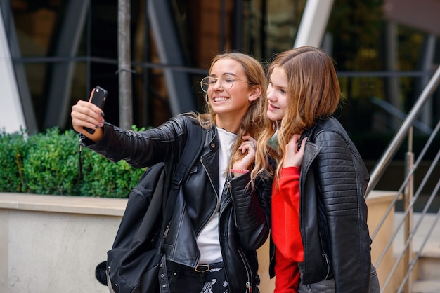 Dos elegantes adolescentes felices con smartphone haciendo selfie cerca de elegante edificio moderno en la calle.