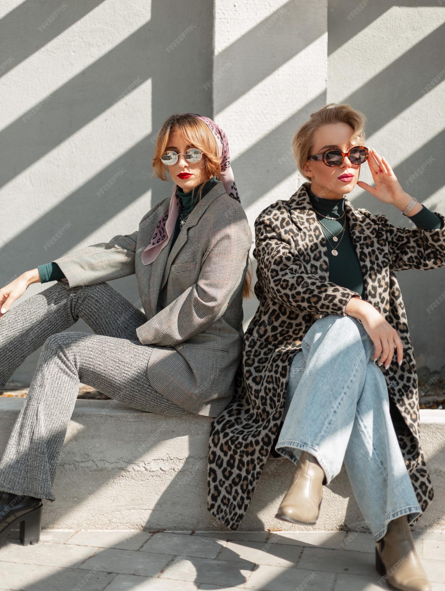 Dos elegantes chicas glamorosas con gafas de sol en ropa retro de moda están sentadas en la calle cerca de un muro de hormigón luz solar traje de estilo urbano