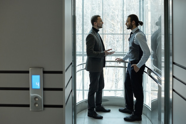 Dos elegantes hombres de negocios discutiendo puntos de trabajo o términos de acuerdo dentro del ascensor del gran centro de negocios