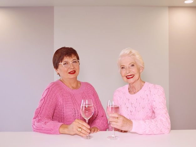 Dos elegantes mujeres de alto nivel en suéteres de color rosa sentado con copas de vino en el bar hablando