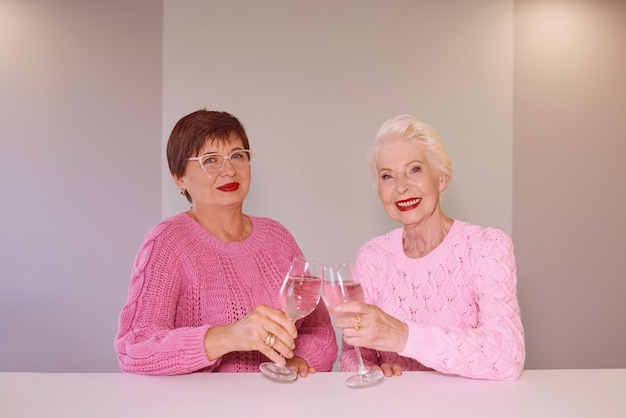 Dos elegantes mujeres de alto nivel en suéteres de color rosa sentado con copas de vino en el bar hablando