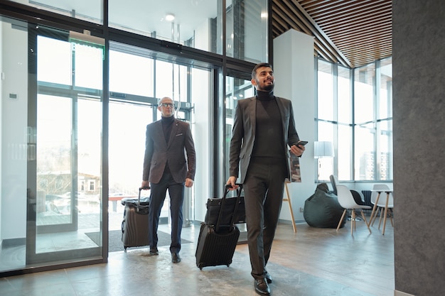 Dos elegantes viajeros de negocios tirando de maletas al entrar en el salón del hotel después de su llegada al país extranjero