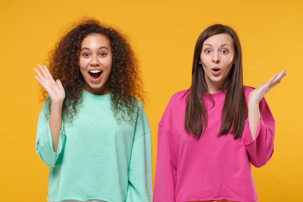 Foto dos emocionadas y asombradas amigas europeas y afroamericanas vestidas de verde rosa posando aisladas en un fondo amarillo anaranjado. concepto de estilo de vida de las personas. simulacros de espacio de copia. extendiendo las manos.