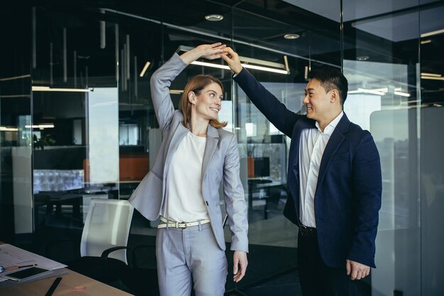 Dos empleados celebran la entrega exitosa del proyecto, un hombre asiático y una mujer rubia bailan juntos en una oficina moderna regocijados y felices colegas