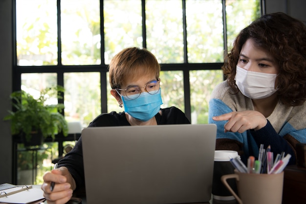 Foto dos empresarias asiáticas en cuarentena y distanciamiento social con máscara quirúrgica mientras trabajaban en la oficina durante la pandemia de coronavirus covid-19