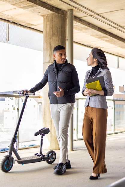 Dos empresarios con scooter eléctrico hablando afuera frente al moderno edificio de negocios