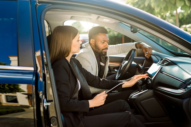 Dos empresarios sonrientes, mujer caucásica y hombre africano sentados juntos en el coche, usando tableta digital y trabajando. Discuta la estrategia comercial mientras conduce a una reunión