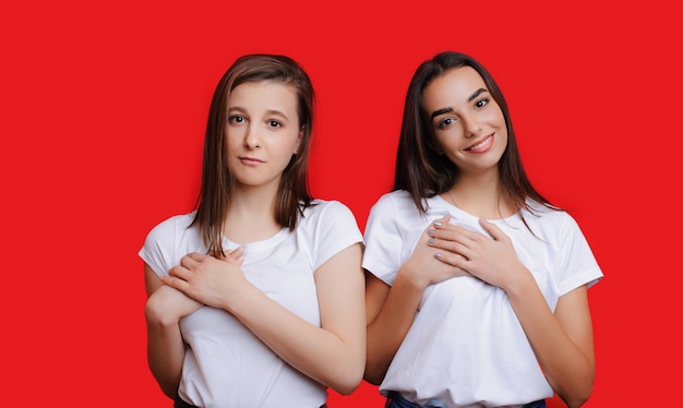 Dos encantadoras chicas caucásicas con cabello negro y hermosa sonrisa tocan sus corazones mientras anuncian algo sobre fondo rojo de espacio libre