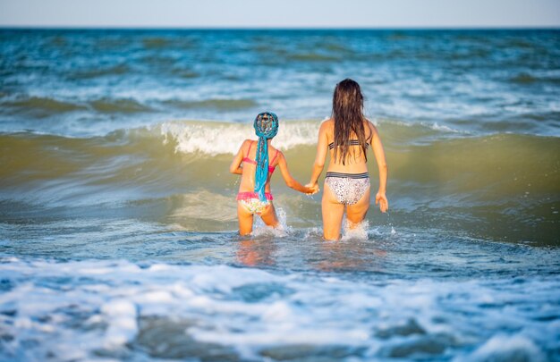 Dos encantadoras hermanas alegres de cabello largo y bronceadas con brillantes trajes de baño de verano se encogen y saltan en olas azules y espumosas en un banco de arena con pequeñas conchas marinas en una cálida tarde soleada de vacaciones