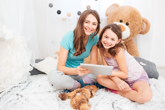 Dos encantadoras hermanas sonrientes sentados y usando la tableta juntos en la habitación de los niños