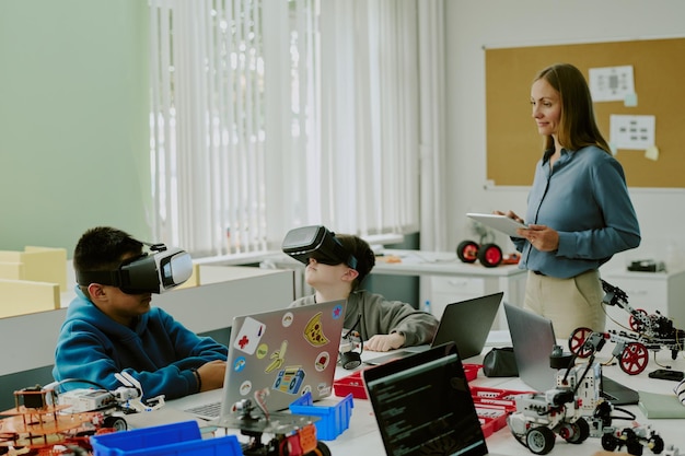 Dos escolares disfrutando de un juego de realidad virtual mientras están sentados en el escritorio con computadoras portátiles y robots maestra