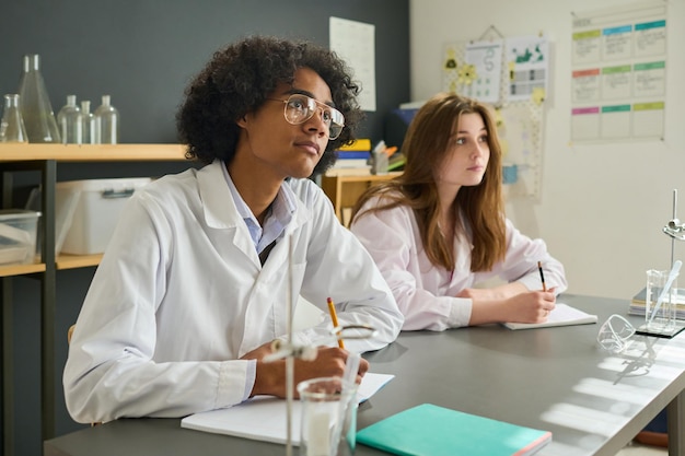Dos estudiantes adolescentes en batas de laboratorio mirando a su maestro mientras lo escuchaban