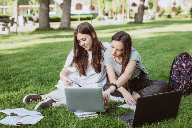 Dos estudiantes están sentadas en el parque en el césped con libros y computadoras portátiles, estudiando y preparándose para los exámenes. Educación a distancia. Enfoque selectivo suave.