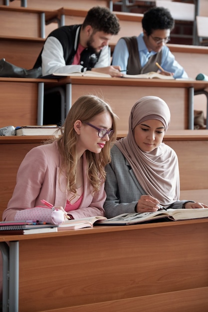 Dos estudiantes interculturales de colegio o universidad sentados en la primera fila junto al escritorio en la sala de conferencias y discutiendo el pasaje en el libro en la lección