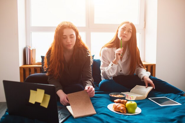 Dos estudiantes pelirrojos estudian en casa o se preparan para los exámenes. Las mujeres jóvenes haciendo los deberes en una cama dormitorio cerca de la ventana. Hay cuadernos, libros de comida, una tableta y una computadora portátil y documentos