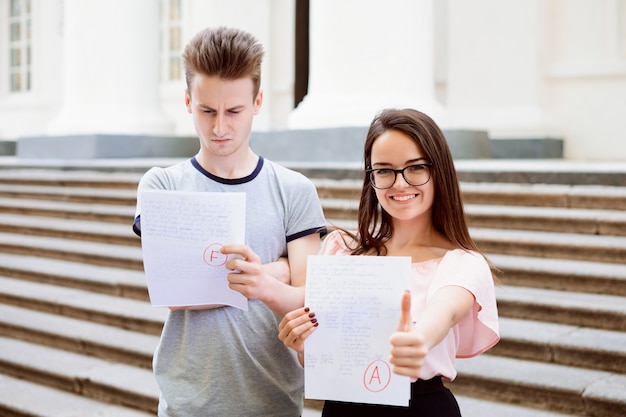 Dos estudiantes con resultados de exámenes. La estudiante feliz ha recibido una excelente calificación A, pero su amigo varón ha reprobado el examen y ha recibido una calificación baja por su trabajo