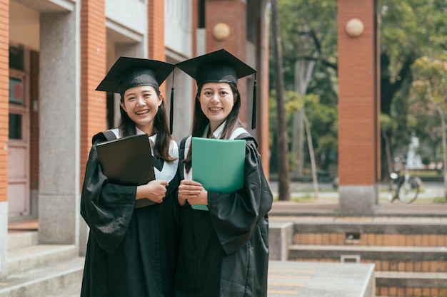 dos estudiantes universitarias asiáticas felices con bata de graduación y gorra sosteniendo libros de texto aprendiendo en la universidad terminaron la educación. mujer amistad cara cámara sonriente confiado encantador lindo alegre alegre