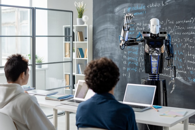 Dos estudiantes varones mirando robot informático de automatización de pie junto a la pizarra con datos técnicos en la lección