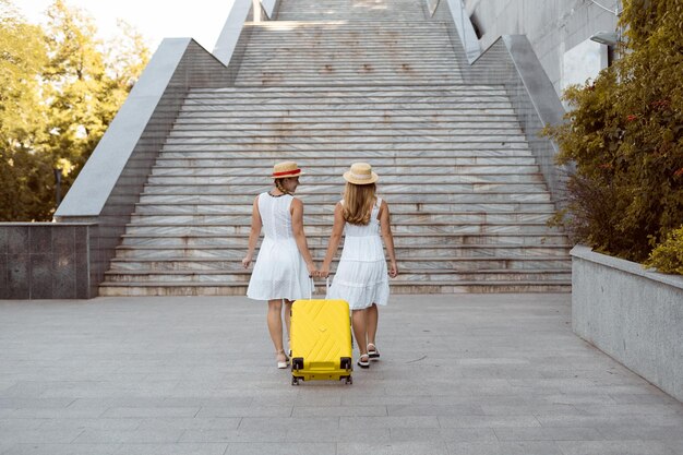 Dos excursionistas gemelos femeninos a la gran escalera. Sacan una maleta amarilla. Copie el espacio.