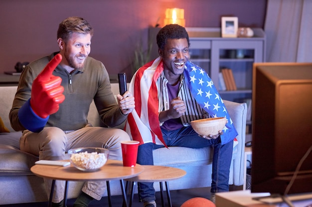 Dos fanáticos de los deportes de cuerpo entero viendo televisión y usando la bandera de EE. UU.