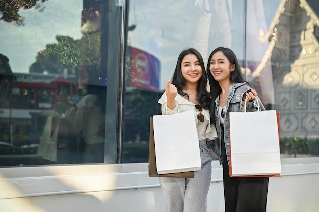 Dos felices compradoras asiáticas paradas afuera del edificio del centro comercial con sus bolsas de compras