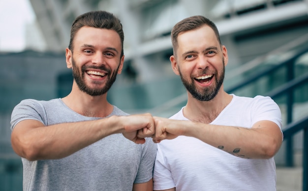 Foto dos felices emocionados mejores amigos barbudos modernos en ropa casual divirtiéndose y charlando juntos al aire libre.