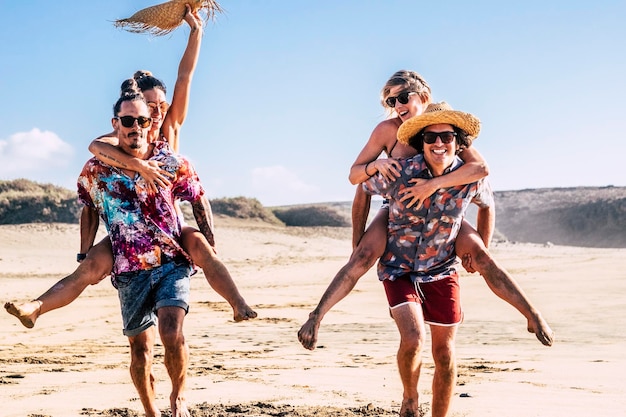 Dos felices jóvenes amigos corren y juegan llevando chicas a cuestas y riéndose mucho juntos en la playa Verano turistas gente vacaciones vacaciones actividad de ocio Hombres y mujeres felices