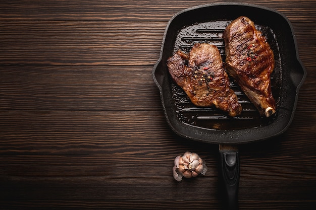 Dos filetes de carne jugosa frita chuletón con condimentos en sartén de parrilla BBQ sobre fondo de madera, vista superior, espacio para texto. Cocinar el concepto de filetes de carne