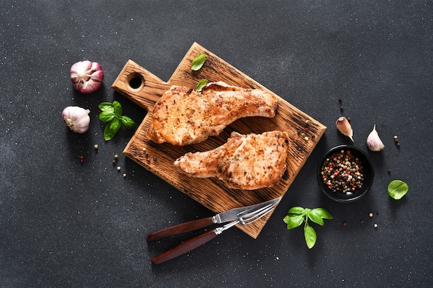 Dos filetes de cerdo a la plancha con ajo y albahaca sobre una tabla de madera sobre un fondo negro con espacio para el texto. Vista desde arriba.