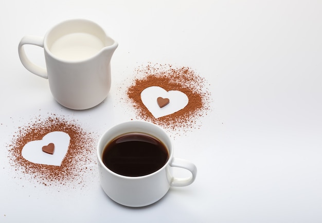 Dos formas de corazón de cacao en polvo, una taza de café con leche y copia espacio sobre fondo blanco.