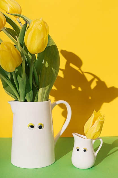 Dos frascos blancos con ojos de juguete grandes y pequeños con ramo de flores de tulipán amarillo sobre un fondo amarillo y verde con sombras nítidas Composición floral de primavera divertida creativa Fondo de pantalla de Pascua
