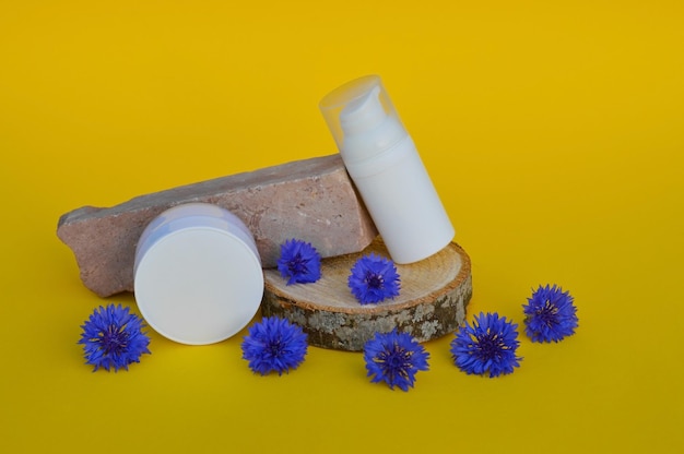 Dos frascos de plástico blanco para cosméticos sobre un podio de madera con flores de maíz azules sobre un fondo amarillo