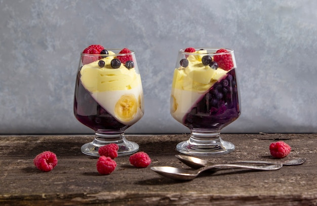 Dos frascos de vidrio con postre frío (panna cotta, gelatina, mousse, pudín) con capas de arándano morado y plátano blanco sobre mesa rústica de madera