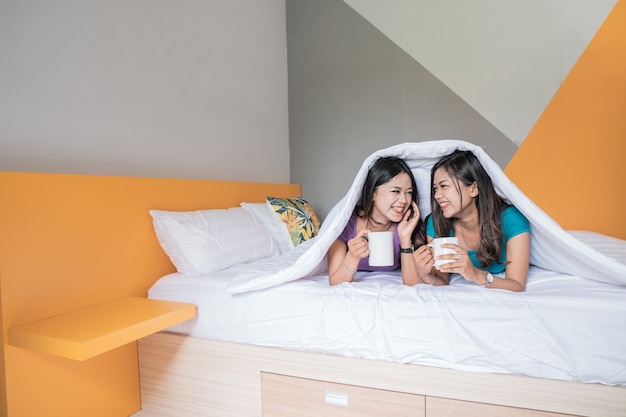 Dos gemelas hermosa mujer asiática sosteniendo una taza con té o café caliente mientras están acostados en la cama juntos