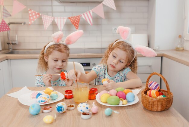 Dos gemelas se preparan para las vacaciones de Pascua en la mesa de la cocina pintando huevos de Pascua