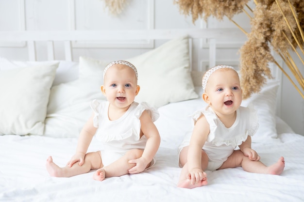 Dos gemelos están sentados en una cama en un dormitorio luminoso y las hermanas pequeñas sonrientes son un bebé con un traje de algodón en la guardería