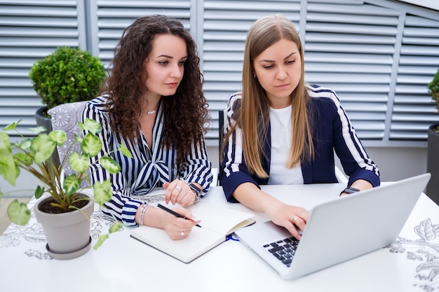 Dos gerentes de mujeres de negocios jóvenes exitosas están sentadas en una mesa con una computadora portátil y un cuaderno trabajando en un nuevo proyecto de desarrollo, las estudiantes escriben un informe sobre el trabajo de su computadora