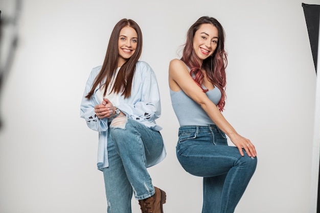 Dos graciosas chicas alegres con ropa casual de denim de moda posan y se divierten en una sesión de fotos con un fondo blanco en el estudio