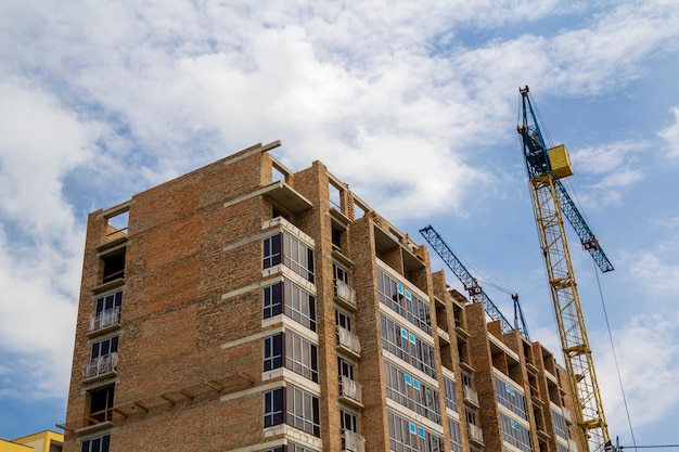 Dos grúas de torre que trabajan en el edificio de ladrillo de gran altura en construcción sobre fondo de espacio de copia de cielo soleado azul. Arquitectura urbana moderna, inversiones, concepto de compra y venta.