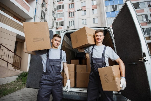 Dos guapos trabajadores con uniformes están de pie frente a la camioneta llena de cajas
