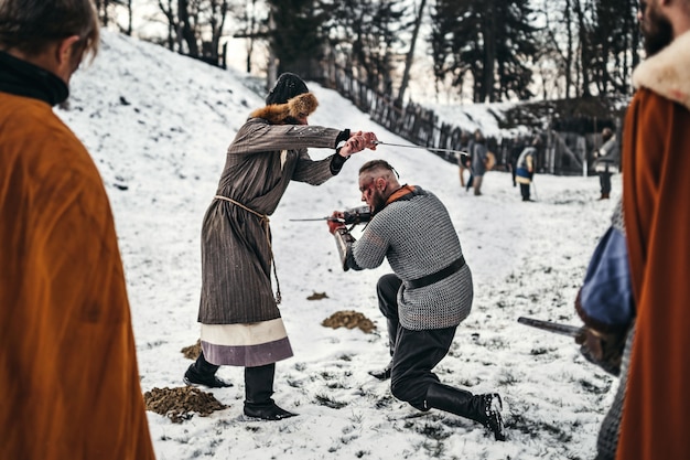 Dos guerreros antiguos en armadura con armas luchando con espadas en la nieve.